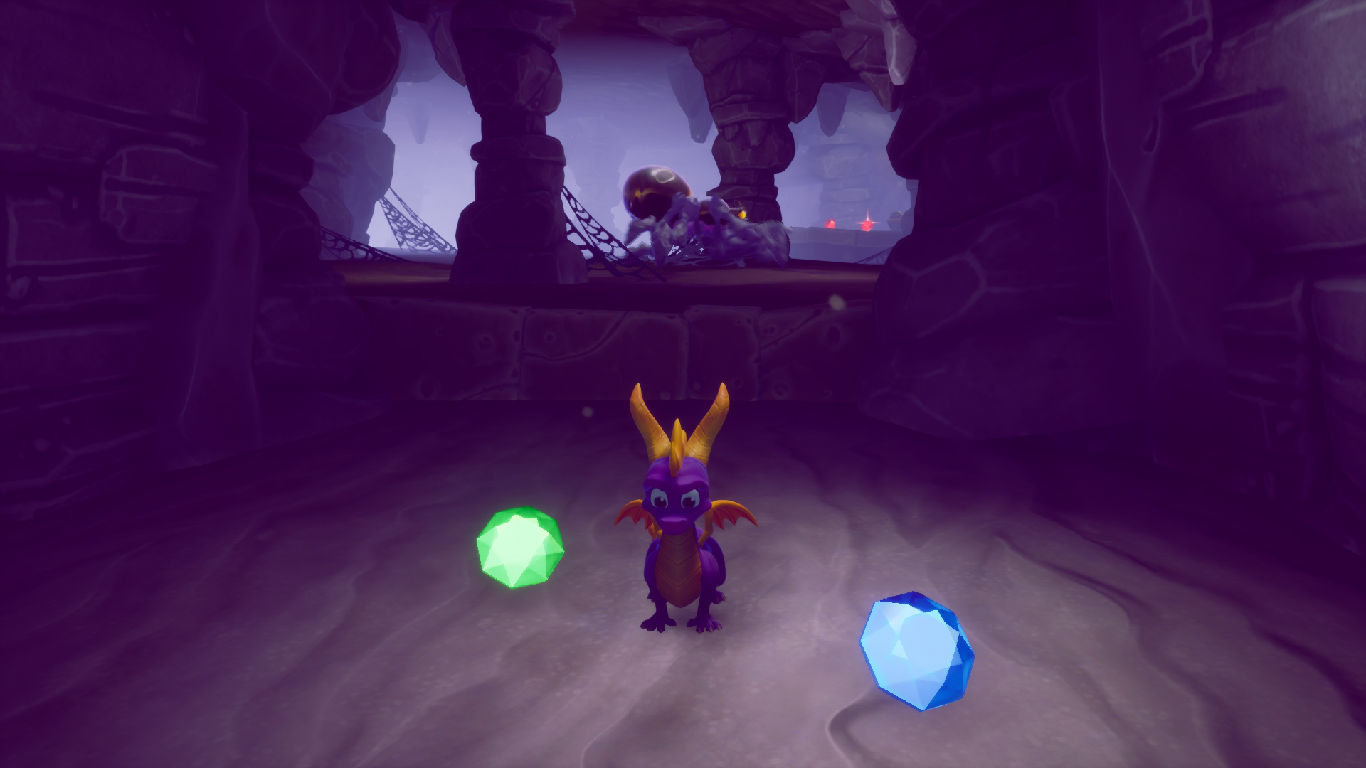 Screenshot from one of the cave levels in Spyro Reignited Trilogy. To the front of the camera is Spyro. Next to him are a green and a blue gem. In the background there is a large spider and to the right of it are two more gems. Those are red.