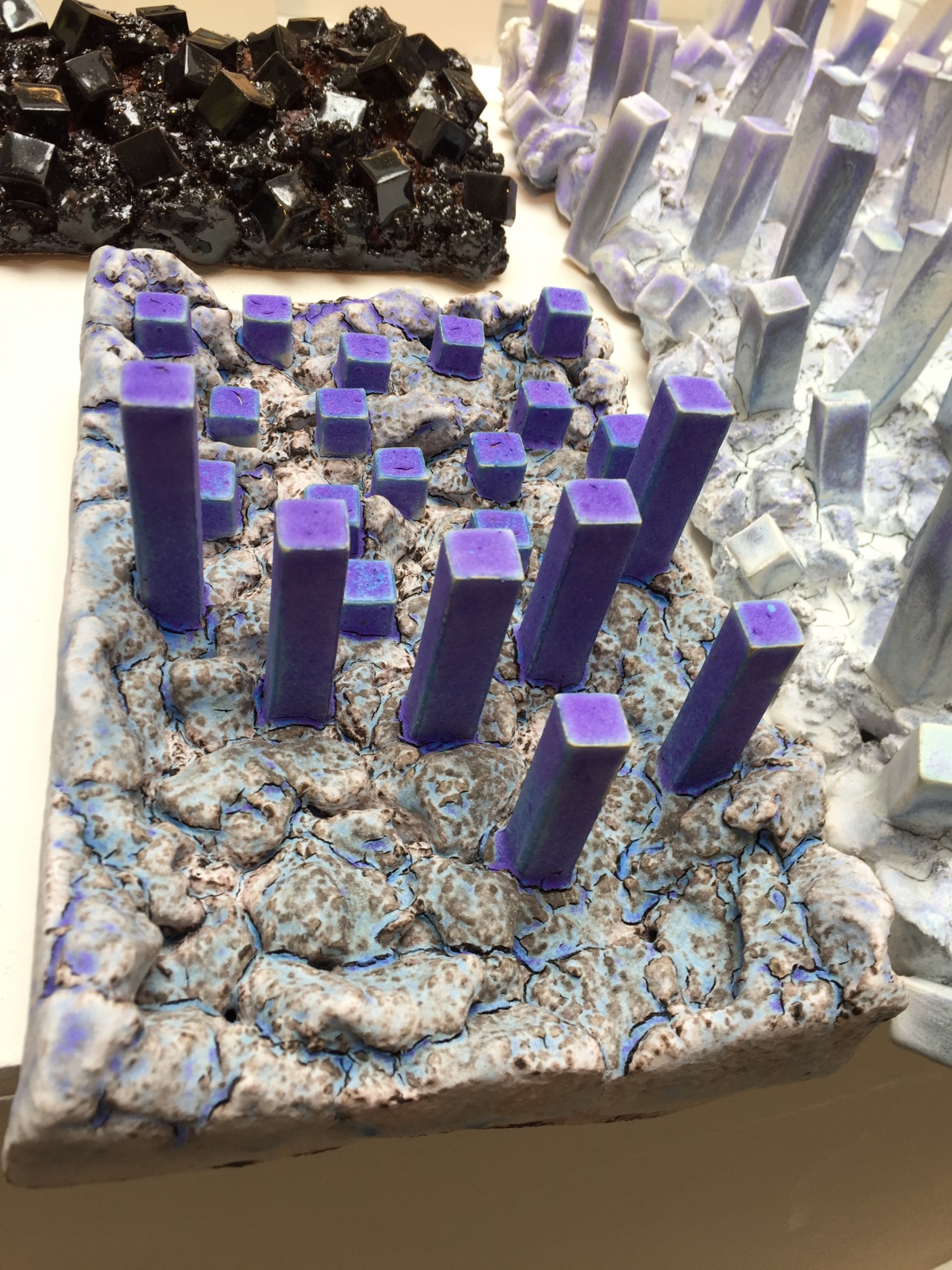 A ceramic sculpture. Captures the image of a flat, rocky ground in white and blues. On top lots of purple parallelepipeds protrude.